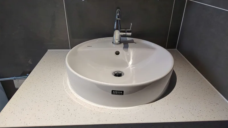Khu lavabo với bồn rửa sứ trắng sáng bóng và tường gạch xám đường ron sáng đều, công trình Bảo Resin tại Đà Nẵng